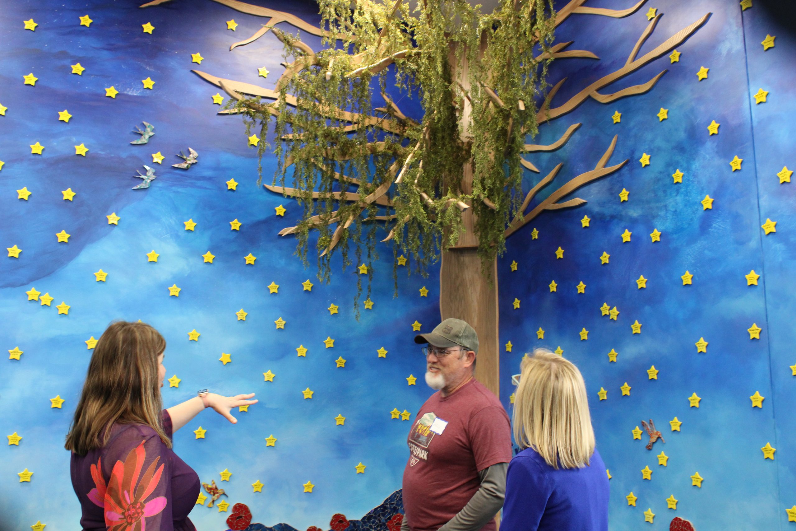 Poppy's Mother Christie Babinski, mural artist Garrick Cline, and Executive Director Summer Kelly in front of the decorative tree with yellow stars and sky mural painted behind them.